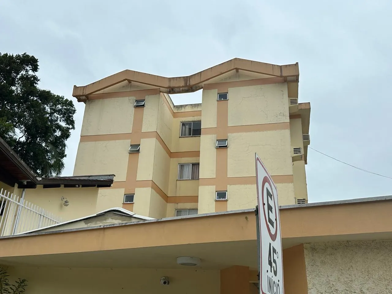 Four-story residential building in yellow and orange tones, with white windows and air conditioners, under a cloudy sky