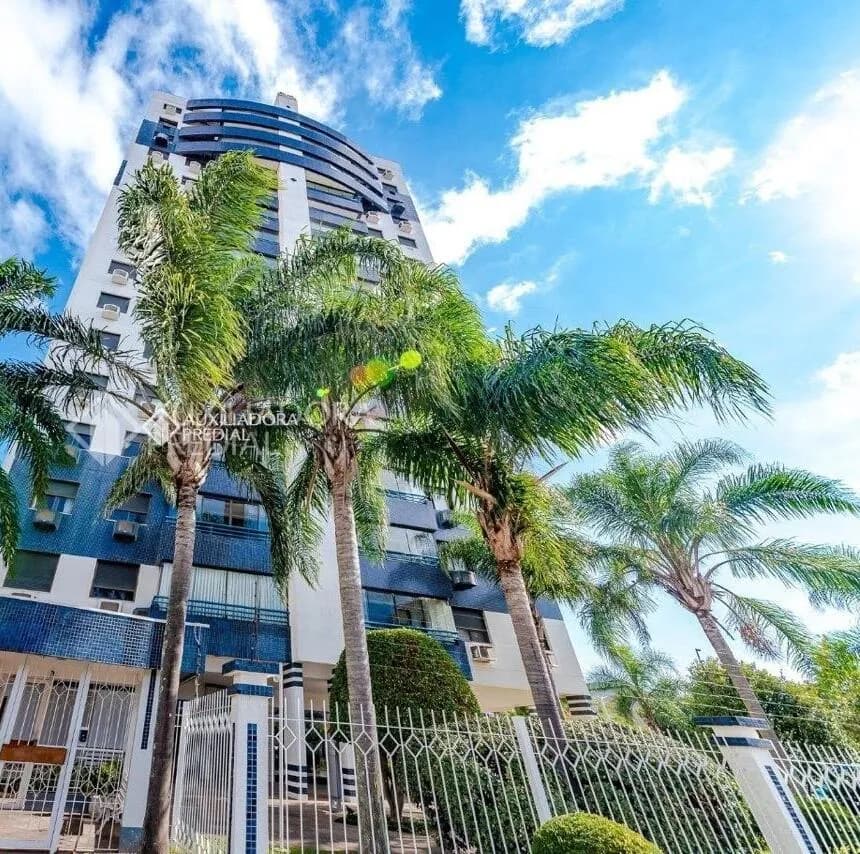 Prédio residencial alto em branco e azul visto de baixo, com palmeiras e gradil branco na frente, sob céu azul com nuvens
