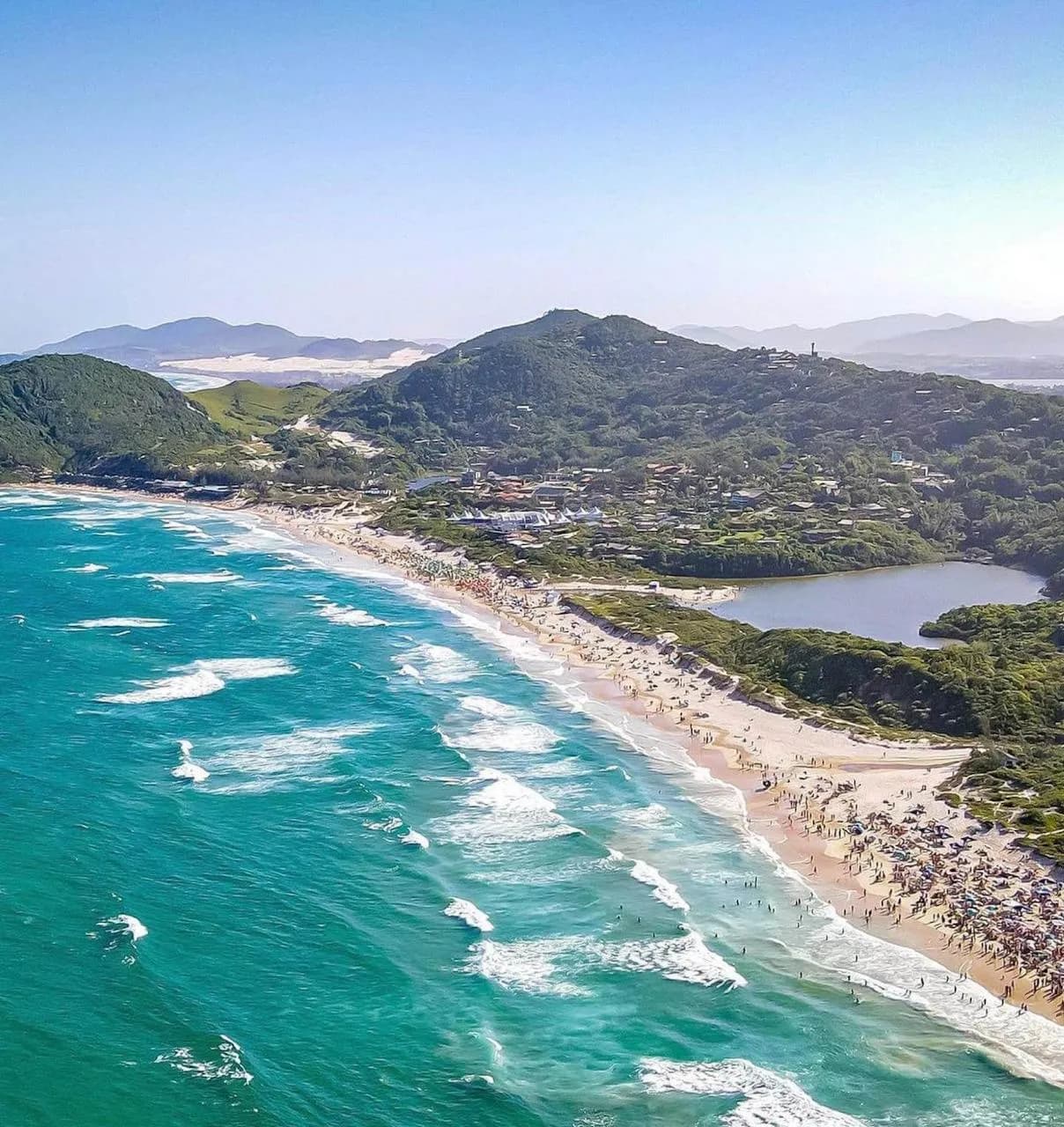 Vista aérea da Praia do Rosa: mar verde-azulado com ondas, a faixa de areia, uma lagoa atrás da praia e os morros cobertos de mata ao fundo