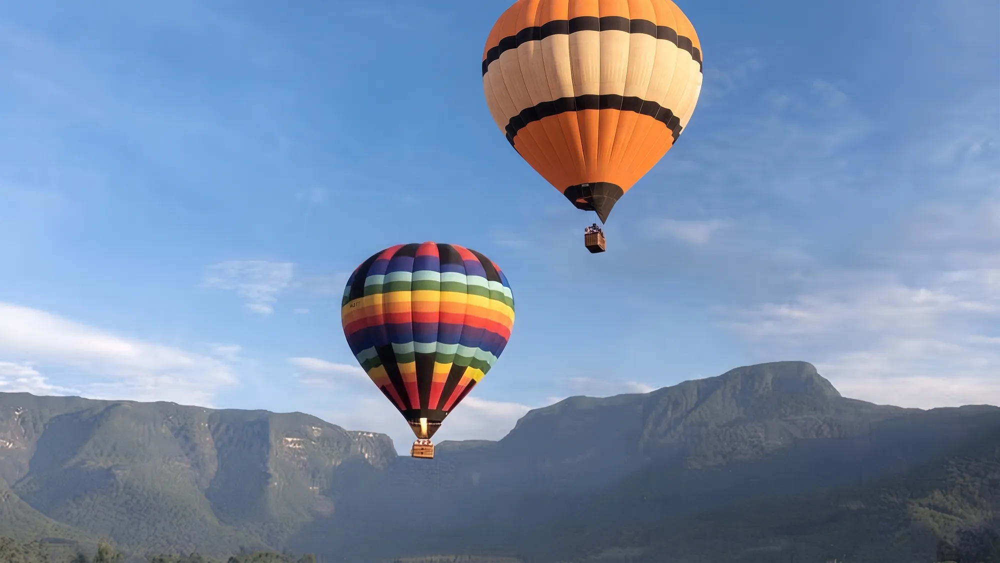 Imagem ilustrativa de dois balões de ar quente, um laranja e um listrado colorido, sobre os paredões da serra sob céu azul