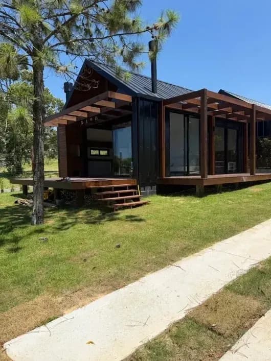 Tiny house na Praia do Rosa: fachada de madeira escura e grandes portas de vidro, deck com escada e pérgola de madeira, gramado e pinheiro à frente